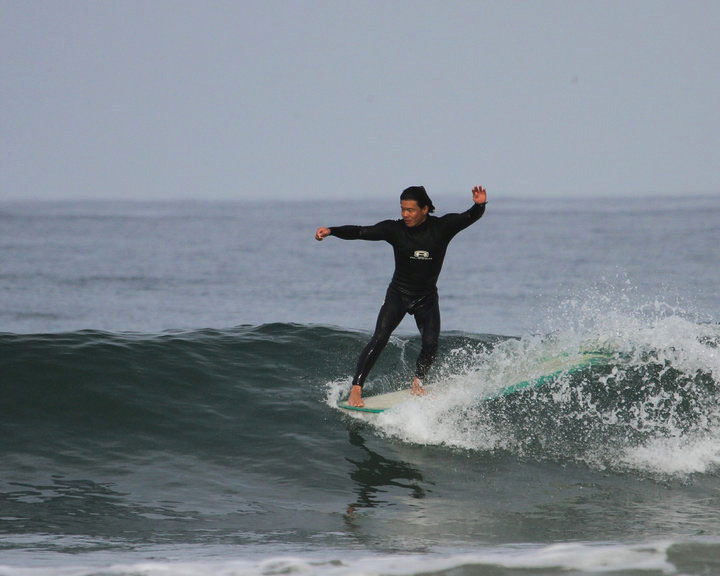 Dale surfing a longboard. He is going right, and his right foot is almost to the tip of his board. His surfboaed is white with green colored rails. The wave is about waist high.